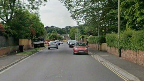 Berrow Road with cars parked either side and houses on the left. Trees and walls with bushes line either side of the road.