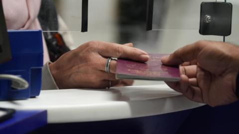 A close-up view of someone handing passing a maroon passport through a small window at a desk. You can see the edge of a blue machine on the left. The hand on the left has a silver ring on the middle finger.