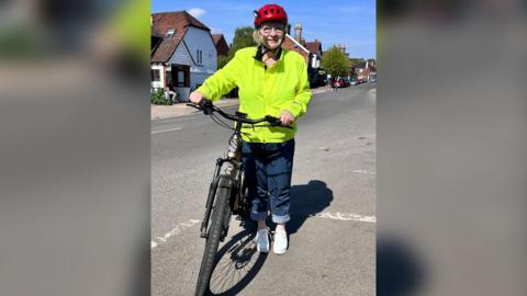 Rosemary Shrager in jeans, a bright yellow high vis jacket and a bike helmet. She is standing on a road next to a bike.