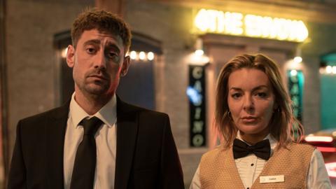 A man and a woman stand side by side with neutral expressions on their faces. The man on the left is wearing a suit and tie, and the woman on the right is wearing a cashier uniform.