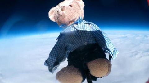 Small, brown bear, wearing school uniform, navy blue shorts and a blue and white checked shirt flying attached to a weather balloon above clouds