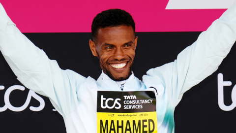 Mahamed Mahamed holding his arms aloft, smiling. He is wearing a white, long-sleeved top. He is standing in front of pink and black banners for the tcs London Marathon. He is wearing a yellow and black sign on his chest saying 'Mahamed'.
