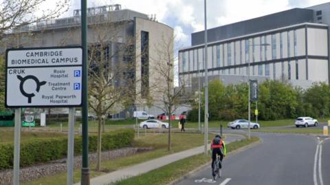 Man cycling on a bike past a roundabout sign with Cambridge Biomedical Campus written on the top of it