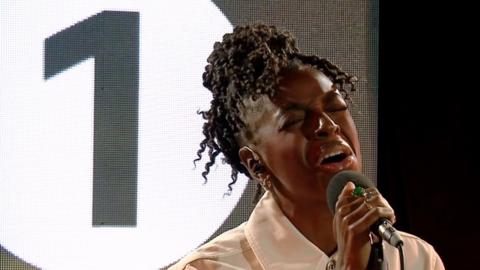 Cat Burns singing in Live Lounge. Wearing White. Radio 1 logo in background