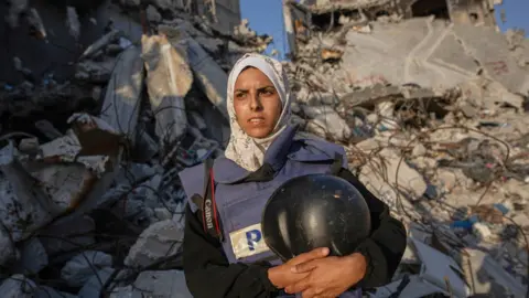 Mariam Dagga wears a flak vest with the word, "press" on the front. She holds her press helmet and has a headscarf. Rubble is seen behind her.