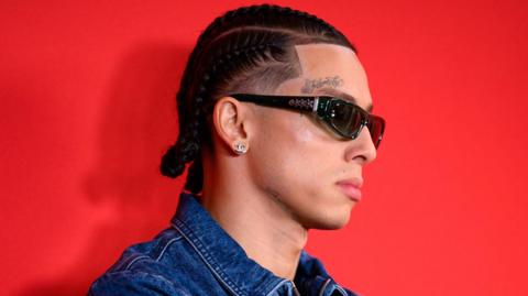 Central Cee  pictured at the GQ awards in 2024. He wears a denim jacket and sunglasses, the tattoo 'family' above his right eye visible in the side-profile shot. He's pictured against a red backdrop. 