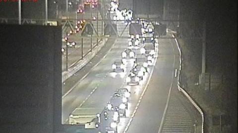 Long tailbacks of cars and lorries on two lanes of the M4 at junction 19 westbound, towards the slip road for the M5. The vehicles all have headlights on. The middle two lanes are empty.