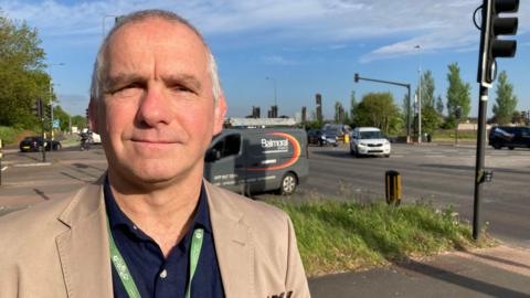 Councillor Sean Rhodes wearing a dark shirt and a tan blazer. He has short grey hair and is clean shaven and is standing beside a road with cars passing by. It is a sunny day and he is smiling at the camera.
