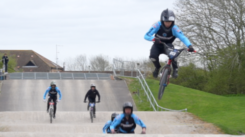 A group of BMX riders hurtling down a track. There are five of them each on their bike in the picture. At the front one of the riders is going over a bump on the track and has been propelled into the air.