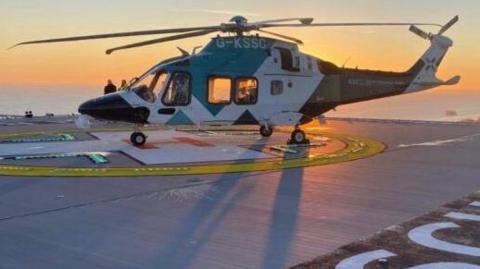 A blue and white helicopter on a helipad, with the letters RSCH painted on the surface. An orange sunset and a view over the city of Brighton can be seen in the background.