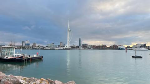 The Spinnaker Tower sits prominently at the back of the photo under clouds and in front of a large area of water that is largely aside from a few small boats