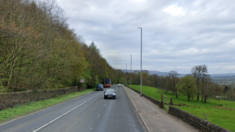 Cars driving down a three-lane road with street lighting. There is fields on the right hand side and trees on the left.