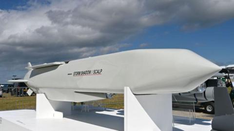 A Storm Shadow cruise missile, which is several metres long and has a white glossy outer layer, as well as a "Storm Shadow/SCALP" logo, pictured on a display stand at an air show in Farnborough 