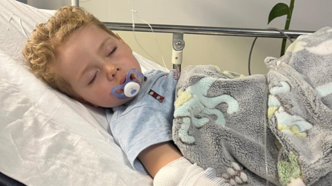 Edison with a dummy in his mouth and asleep on a hospital bed. He has a fluffy blanket on top of him.