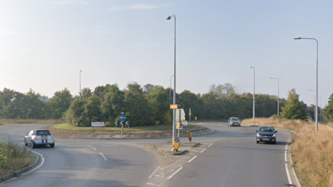 A Google streetview image of a roundabout with trees on it. There are three cars being driven around it. There are lampposts dotted around the roundabout and dry grass. It is sunny and the picture has clearly been taken in summertime.