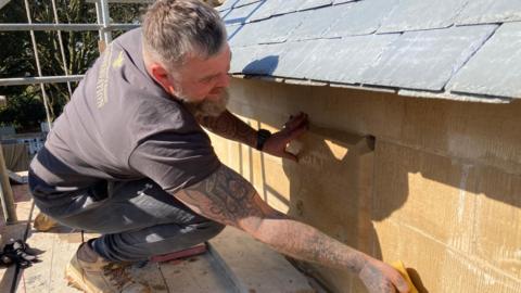 a tattooed man wearing a t-shirt sponges down sandstone while standing on scaffolding at the church. You can faintly see an engraving on one fo the stones saying High Toynton 2025