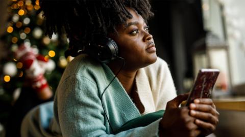 Someone lying down on the floor in a jumper wearing headphones and holding a phone. There is a Christmas tree in the background with lights on.