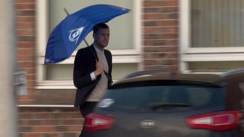 A man holding a blue umbrella walking between a car and a building.