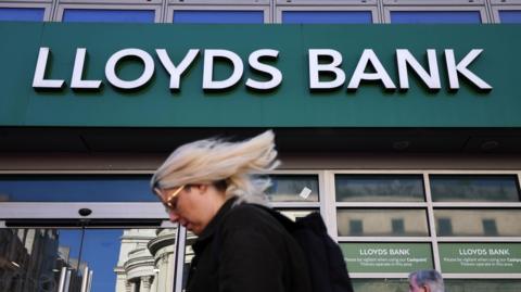 The Lloyds Bank logo on the outside of a high street bank. In the foreground a woman with blonde hair walks past