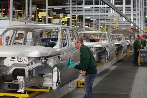A factory line of shells of cars- a man wearing a green jumper, safety goggles and a green glove is standing sideways working on one of the cars. Colleagues can be seen in the background also wearing green jumpers. 