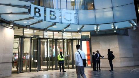 A person wearing a white shirt and carrying a shoulder bag walks towards the entrance doors at BBC Broadcasting House in London on Monday, with the BBC logo in view.