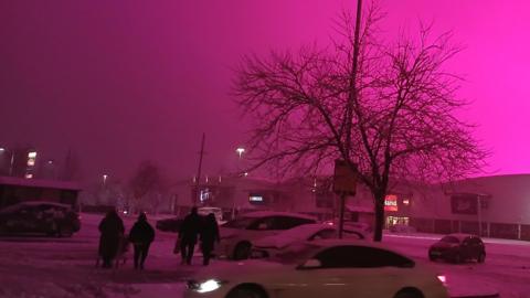 A magenta sky above a retail park car park. The car park is mostly empty, save for a few cars, and is covered in snow. A group of four people is walking across the car park