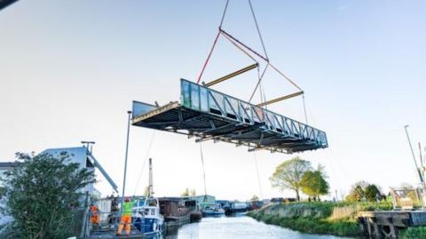Weel Bridge being hoisted from its position on the River Hull. The bridge platform is in mid-air with a person in hi-vis clothing standing nearby. There are a number of boats moored on the waterway.