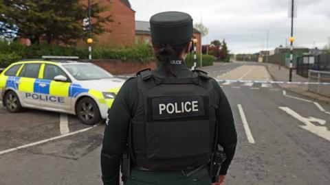 A female police officer stands in front of a police car covered with green and blue patches. She is wearing a green uniform and a vest saying 'police'