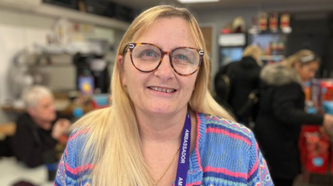 A woman with long blonde hair and round glasses smiles at the camera. There are a number of people, blurred out, behind her.