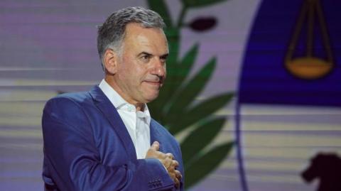 Yamandú Orsi, wearing a blue suit and a white shirt, places his right hand on his heart in a gesture to thank his supporters as early results come in on election night. 