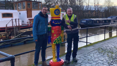 Two men stand either side of the colourful nutcracker statue on the riverside.