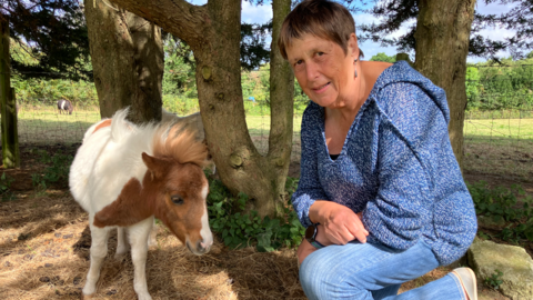 Lucy is pictured next to her owner, Jean. She has short hair and is wearing a blue jumper with blue jeans. The foal is white and brown in colour. They are both stood in a fenced off area surrounded by trees and grassland.