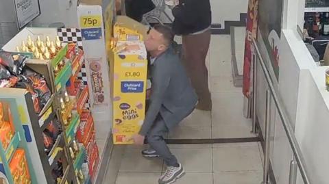 Ashley Daley stealing a pile of mini eggs from a Tesco. He is wearing a grey jacket and trousers.