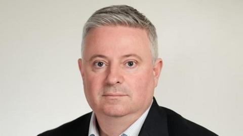 Paul Scott looking at the camera with a serious expression. He is wearing a black suit jacket and a white shirt without a tie. He has short grey hair.