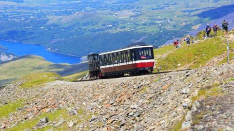 Train carriage seen from the side of mountain as it climbs up, with lake visible in the distance and rocks and grass alongside the tracks. The train is made up of one carriage painted red, white and black and hikers are seen walking on the mountain a little further up