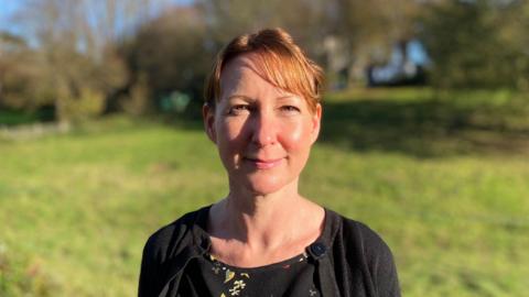 Di Matthews from Public Health, with light brown hair tied up away from her face looking at the camera with a black cardigan and a black floral shirt on standing in front of a blurry field.