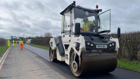 A man dressed in a hi-vis suit sits inside the cabin of a road roller flattening a new road surface. Only half the road has been completed, the other half still has a patchy old road surface.