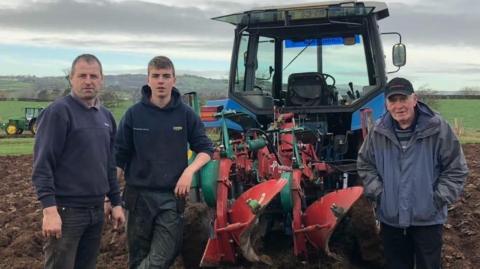 Three men stand in front of a blue tractor with plouging equipment attached. They are in a field, with grey skies and hills behind. There is another tractor in the distance.