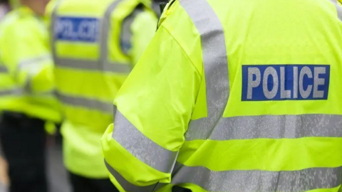 Three police officers are standing with their backs towards the camera, all of them are wearing a bright yellow hi-vis jacket which says police on the back. 