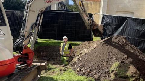 A man wearing a yellow hi-vis tabard and yellow hi-vis trousers stands in a hole which has been dug up by a digger on a patch of grass on a sunny day. The man is smiling. The digger is positioned on one side of the hole, its arm is going over the hole and its bucket is hovering above a large mound of dirt. There is construction fencing in the background and buildings behind.