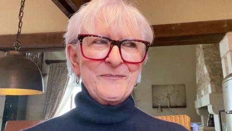 Linda Rands smiles mischievously in a living room. She has short grey hair and wears red glasses.