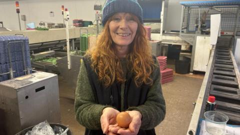 Alison Greene pictured in the processing unit with a conveyer belt running next to her. She is wearing a green jumper with a black woolly gilet and a blue woolly hat. She has red hair and is holding the brown egg in front of her.