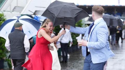 A woman in a red sleeveless dress struggles with her umbrella, while a man in a light blue suit tries to steady it