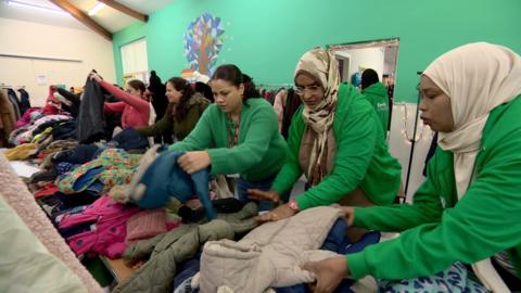 Three women wearing matching green hoodies are in the foreground, arranging children's coats on a table as other women look through coats hanging on rails in the background