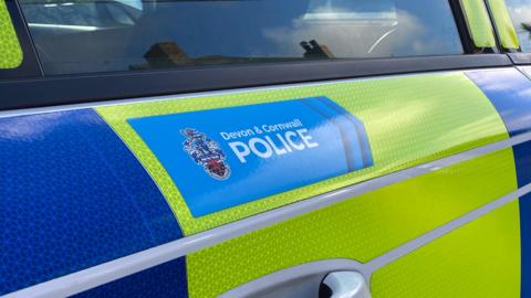 A photo of a blue sticker that reads Devon and Cornwall Police on the side of a yellow and blue police car.