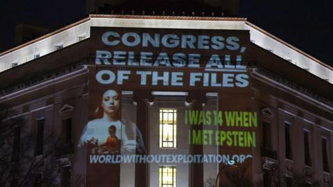 Wording is projected at nighttime onto a building that includes the phrase "Congress, release all files" in capital letters and a white font above a photo of a woman with long brown hair. The words "I was 14 when I met Epstein" are also seen in all capital letters and a green font
