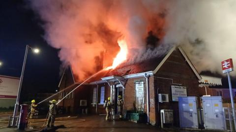 Flames coming out of the roof of a restaurant. Firefighters on the ground are aiming hoses of water at the roof.