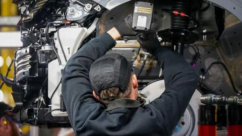 A car mechanic photographed from behind from the shoulders up working underneath the wheel arch of a silver car chassis. He is wearing a dark jumper and a black hat.