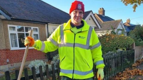 A man wearing a fluorescent jacket and red hat and a broom handle stands in front of a bungalow.