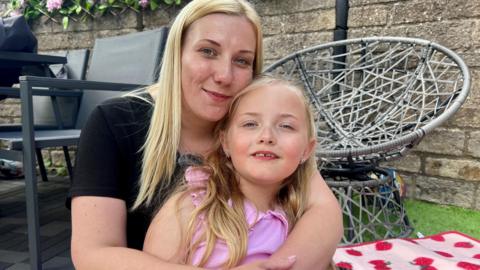 A woman with blonde hair and a black T-shirt sitting on a rug on the grass with her arms around a young girl with blonde hair and wearing a pink top. Both are smiling at the camera and there's a patio chair behind them.